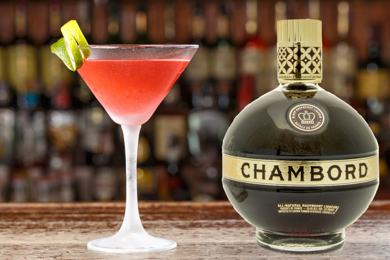 A bottle of Chambord on a bar counter next to a Cosmopolitan cocktail