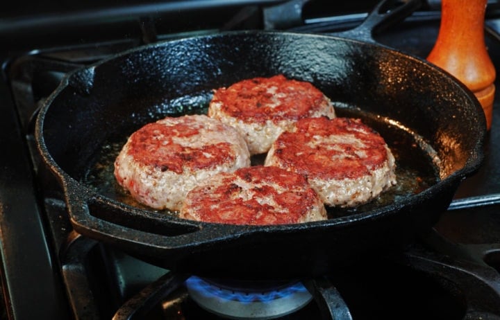Well crusted hamburger patties nearing completion in cast-iron skillet over gas flame with wood pepper mill in the background