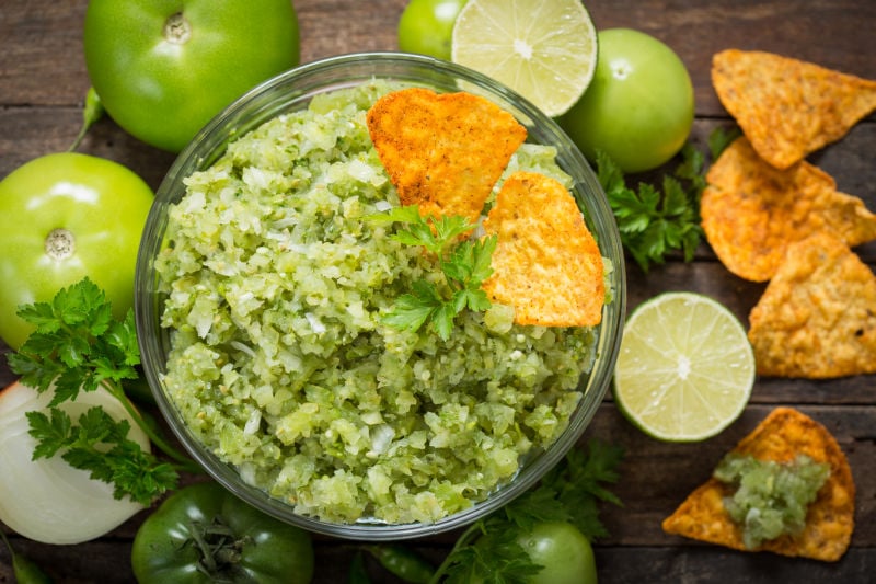 Salsa verde in a dish surrounded by tortilla chips and fresh tomatillos