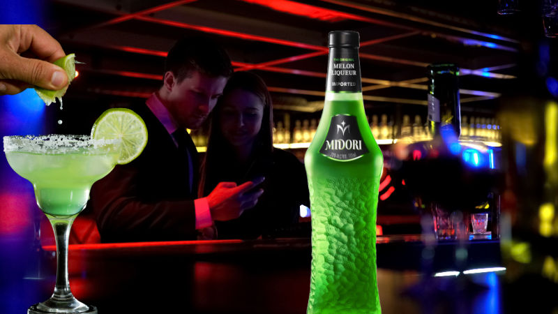 A bottle of Midori on a bar counter next to a cocktail