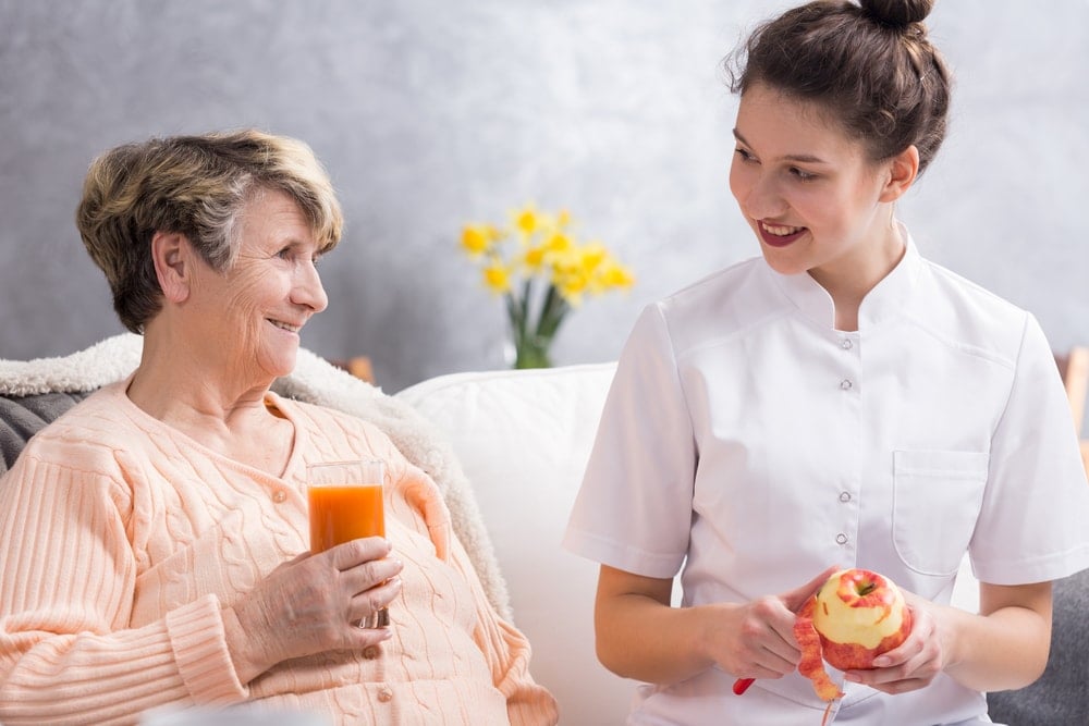 senior woman drinking apple juice while caregiver peels a red apple