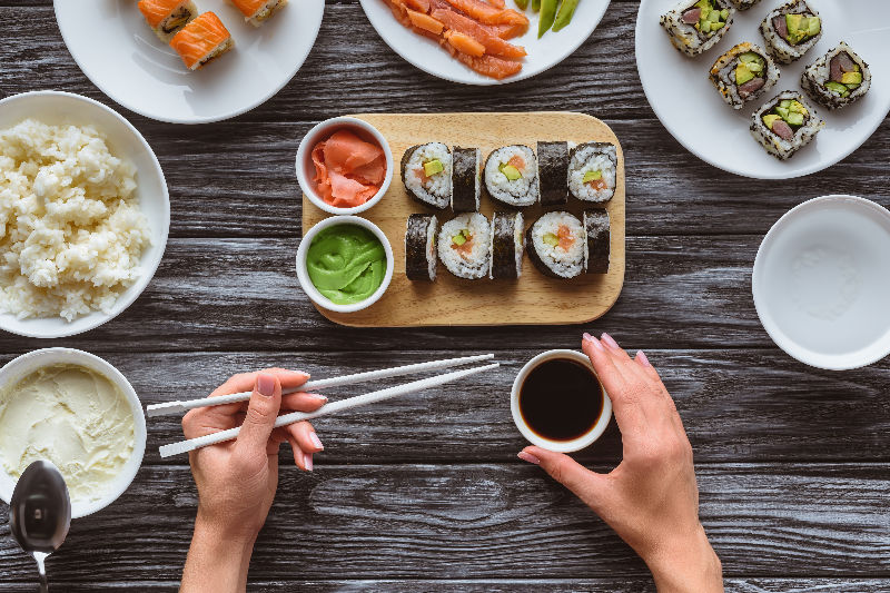 A selection of sushi rolls on a table and a lady holding chopsticks