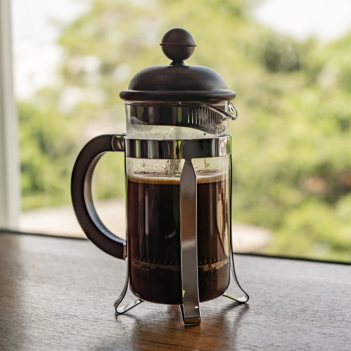 french press containing coffee on windowsill trees outside window in background