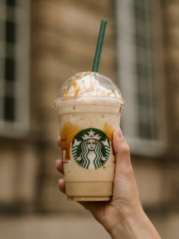 Hand Holding Up Cup Of Starbucks Frappuccino Stone Building In Background 360x480