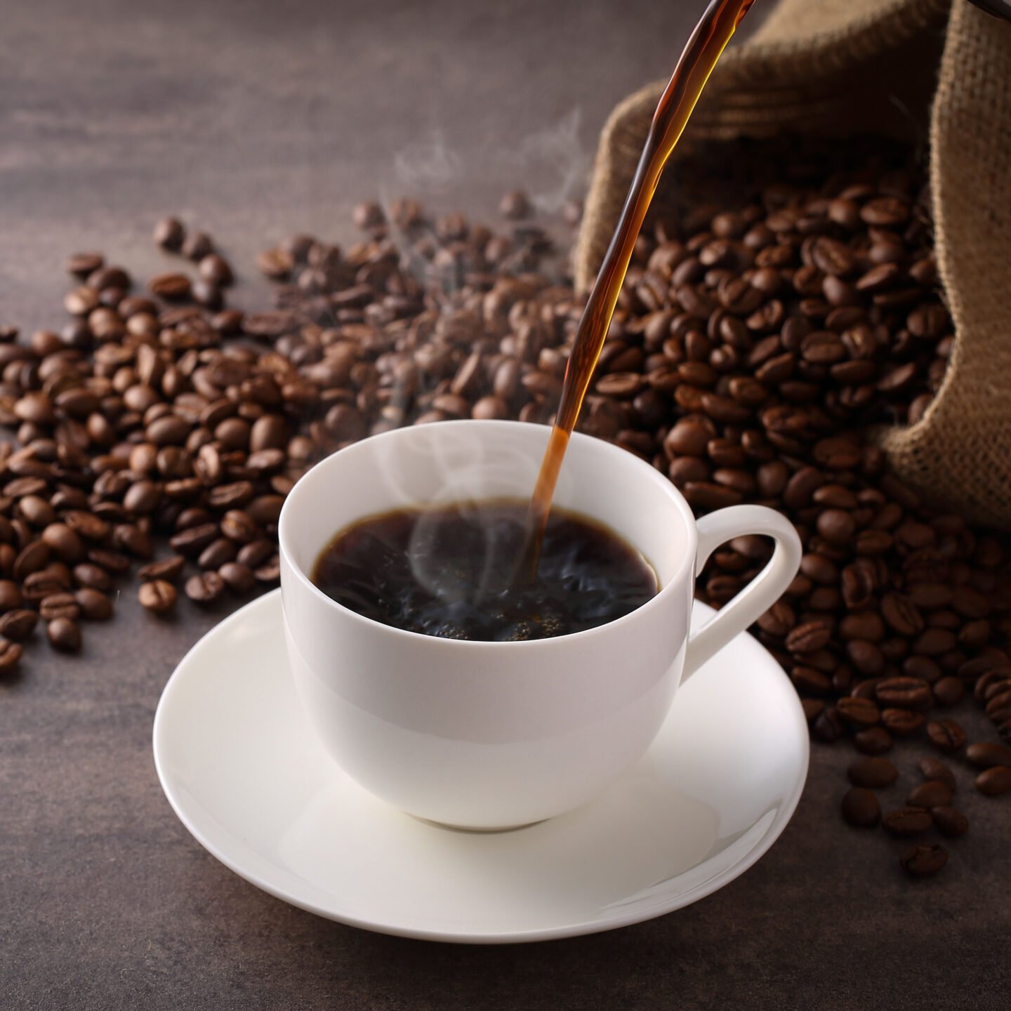 pouring freshly brewed black coffee into white mug on saucer whole beans in background