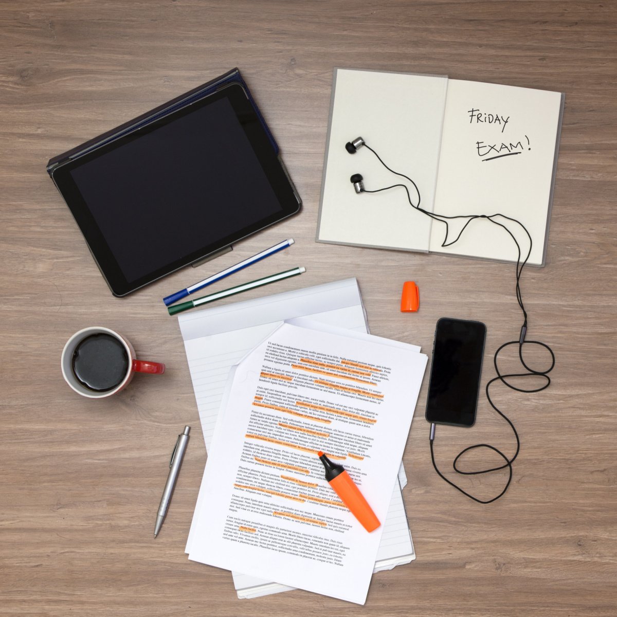 coffee in red ceramic mug beside highlighted papers pens and gadgets on wooden table