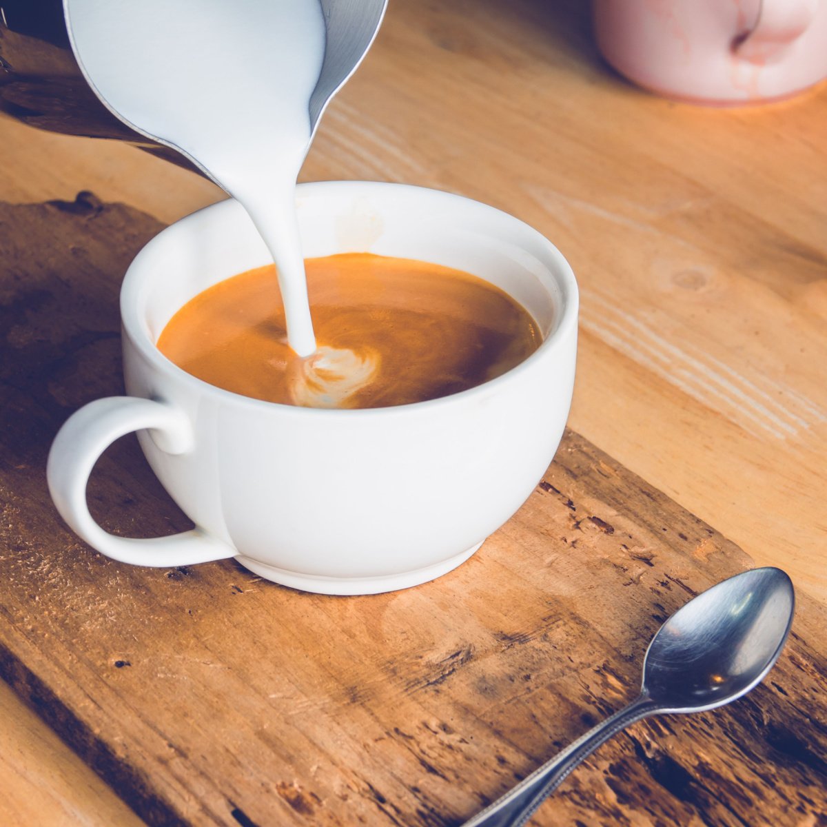 pouring milk or cream from stainless jug into cup of coffee on wooden table