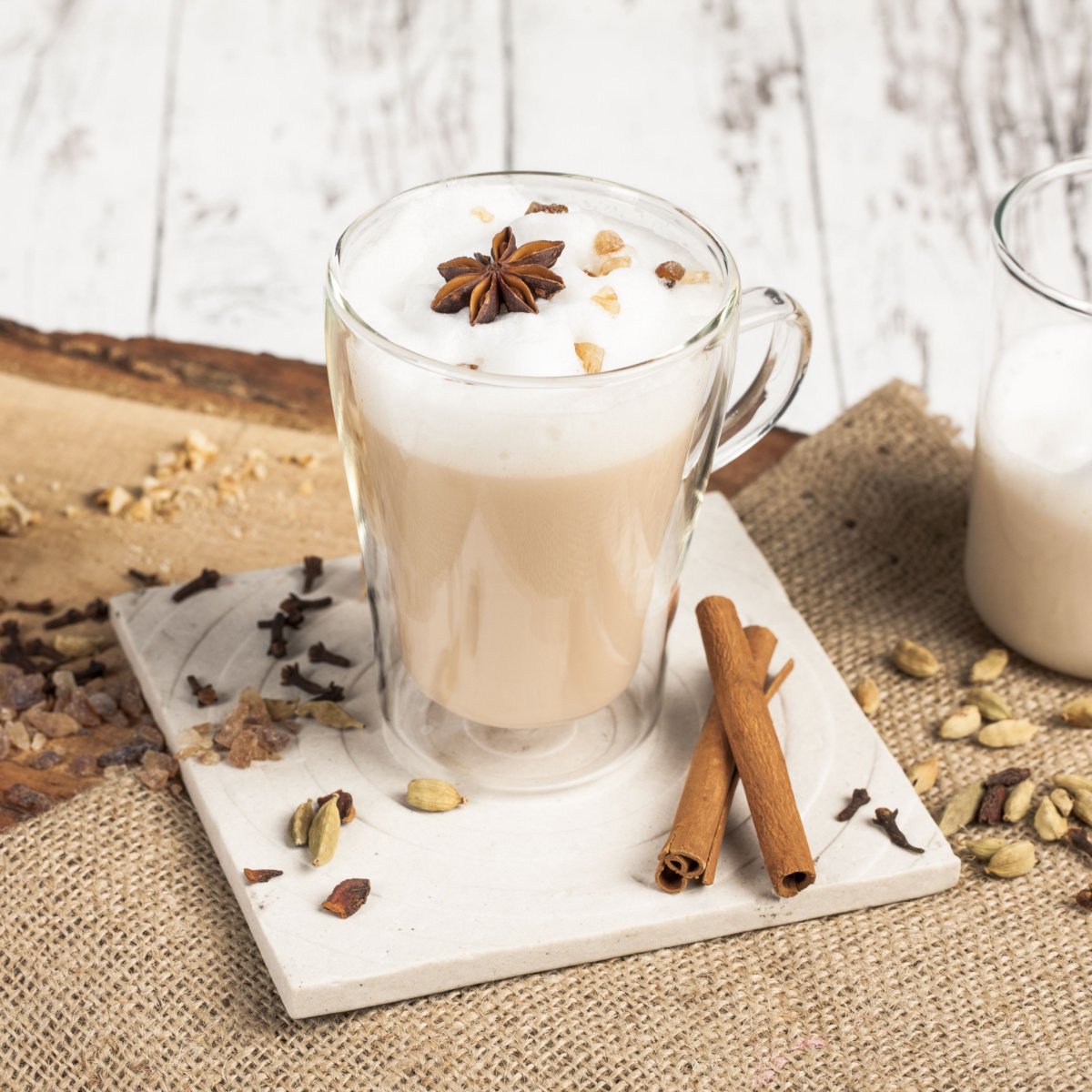 chai latte in tall glass mug beside jug of milk surrounded by spices