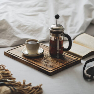 coffee in french press and mug on wooden tray beside a bed and open book