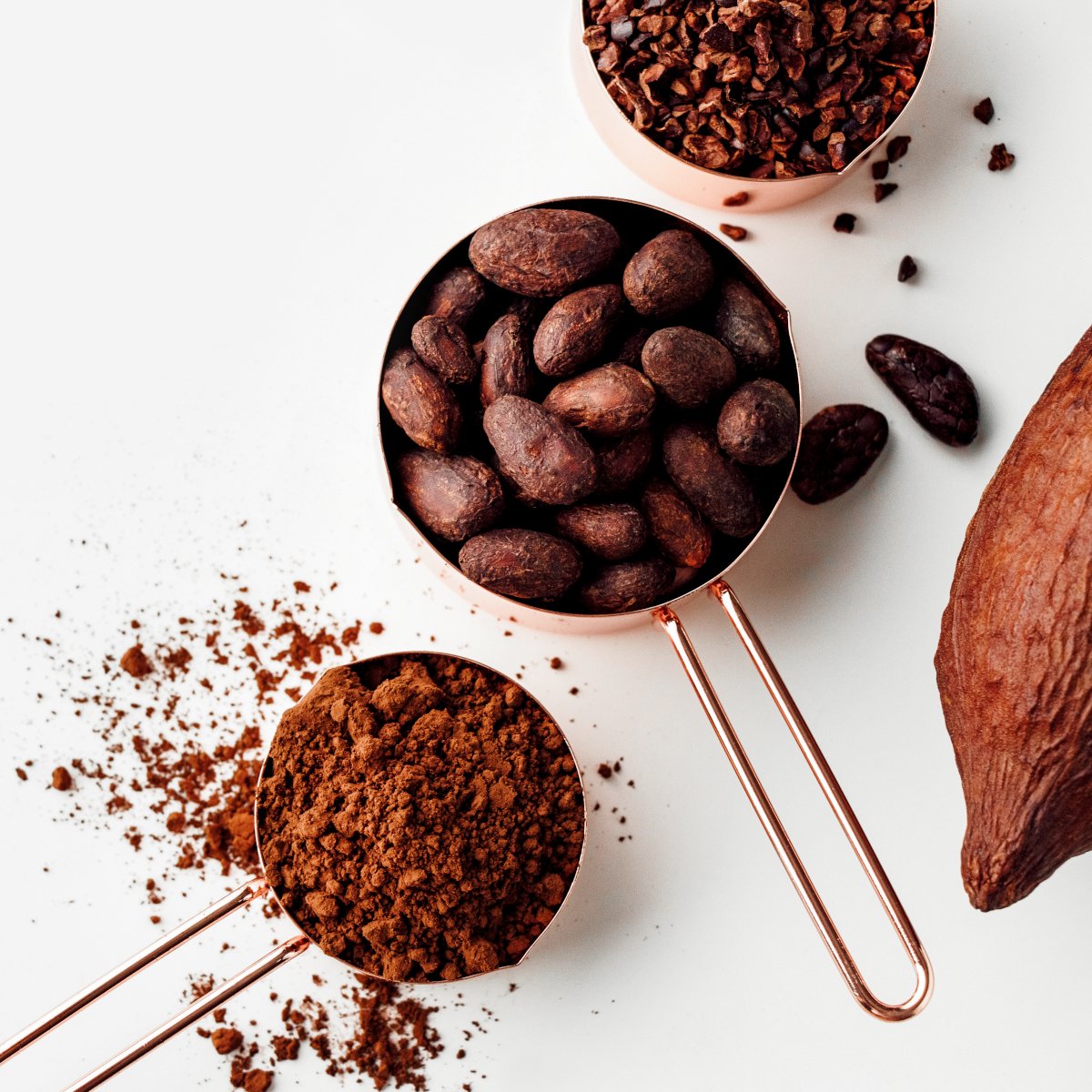 rose-gold-measuring-cups-with-cocoa-beans-cacao-nips-cocoa-powder-and-cocoa-pods-on-a-white-background