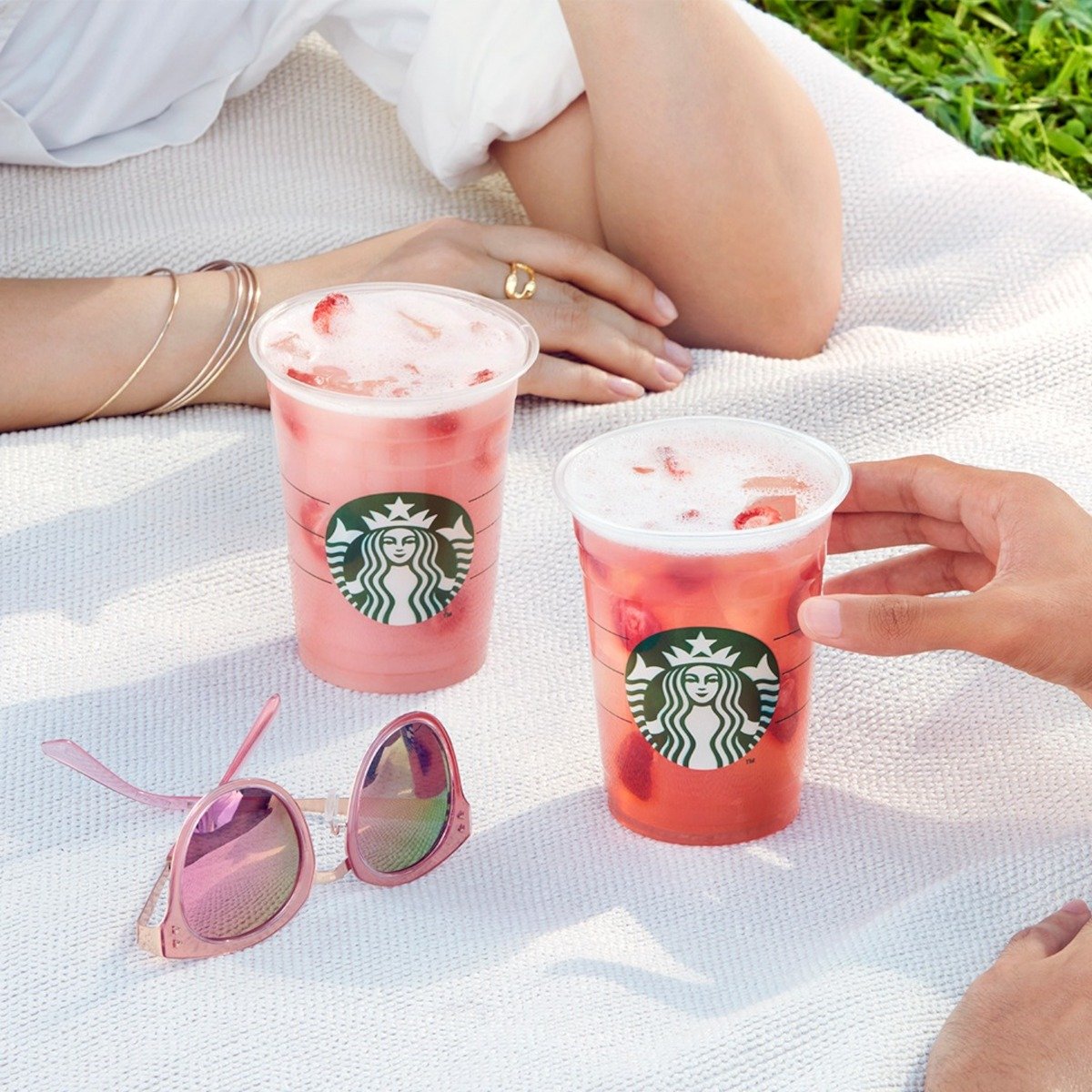 two pink starbucks drinks beside a pair of sunglasses on a picnic blanket