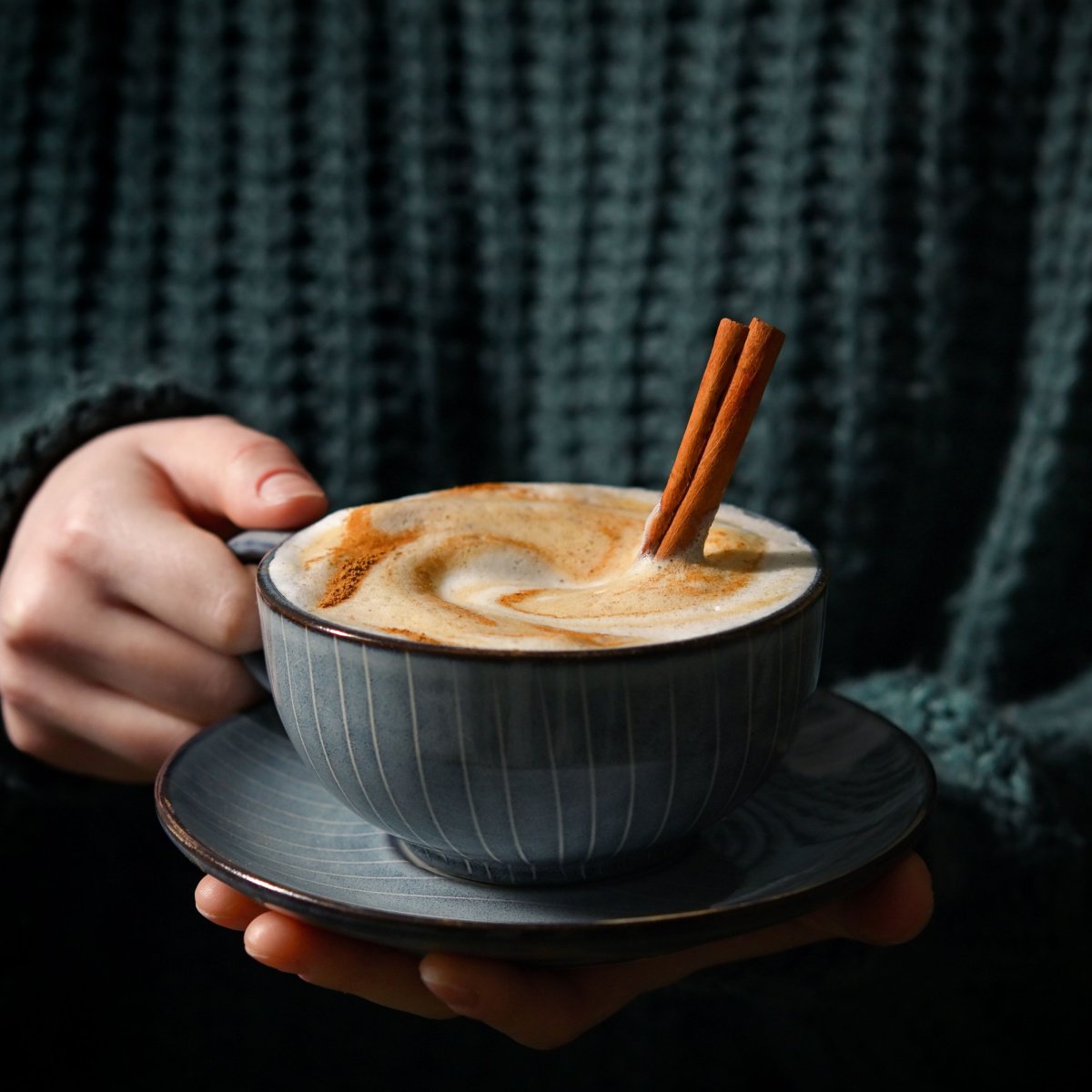 hand holding cup of coffee on saucer topped with cinammon and with cinnamon sticks sticking out
