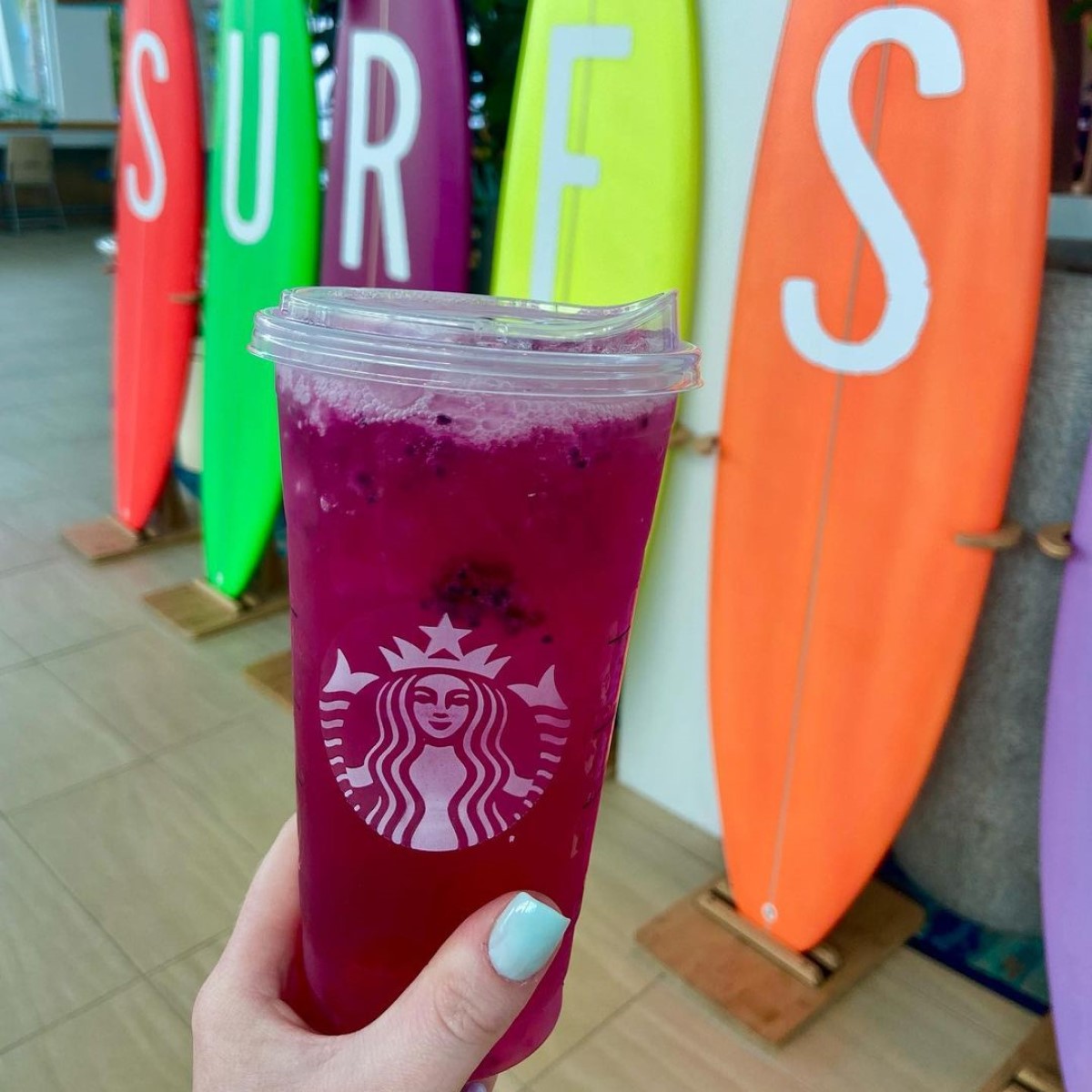 hand holding cup of starbucks mango dragonfruit refresher with surfboard in background