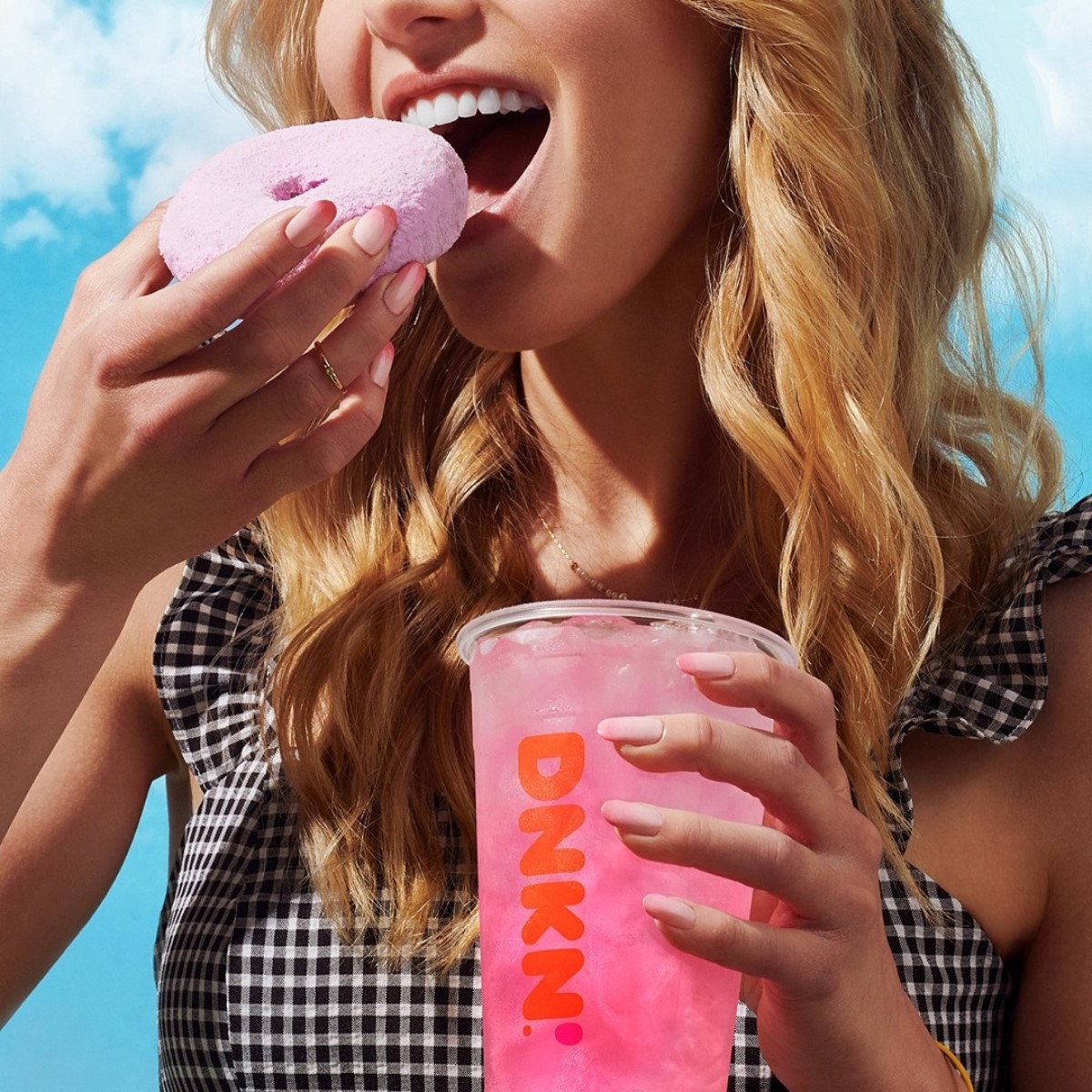 woman holding cup of strawberry flavored lemonade dunkin refresher while biting into donut