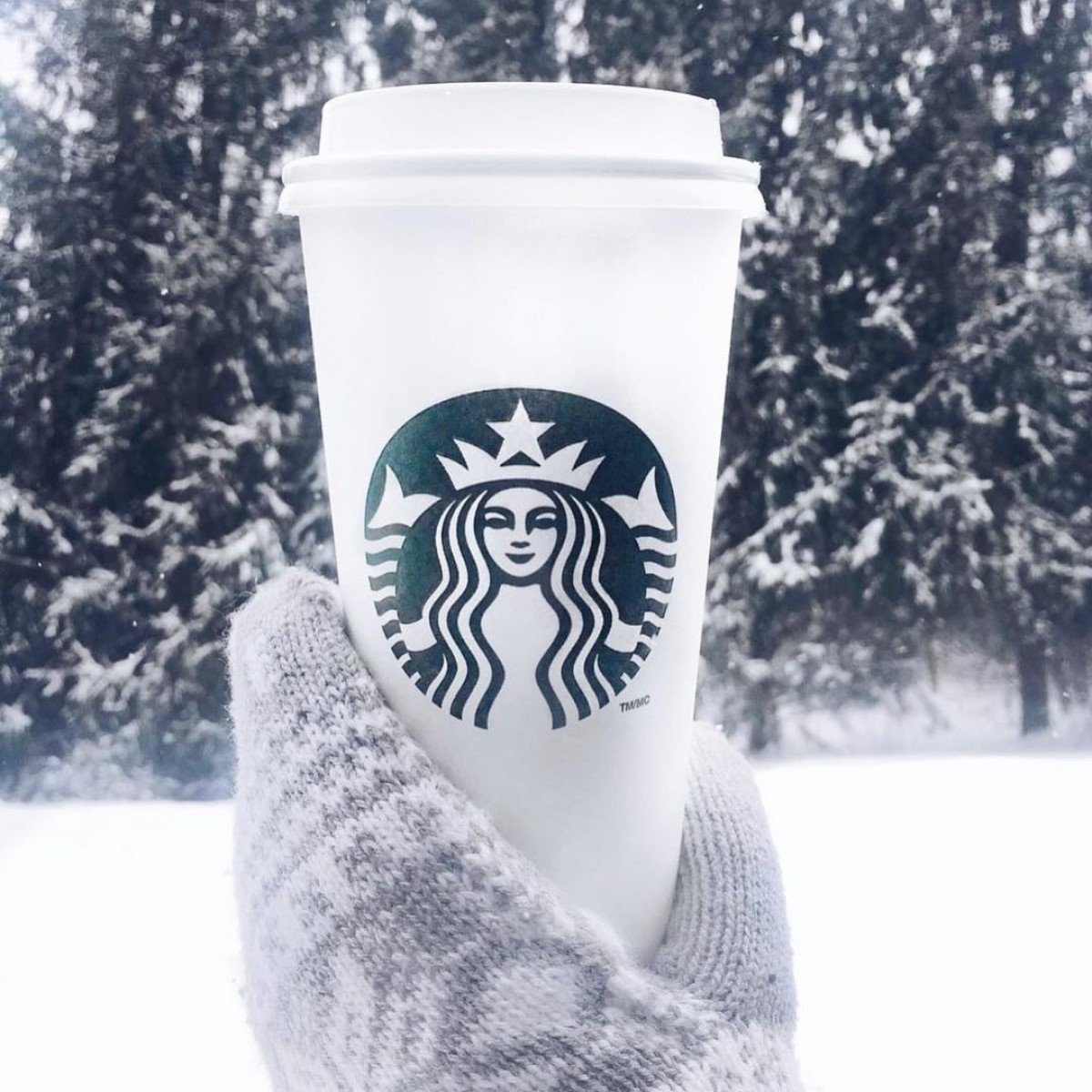 gloved hand holding up white starbucks paper cup in the snow