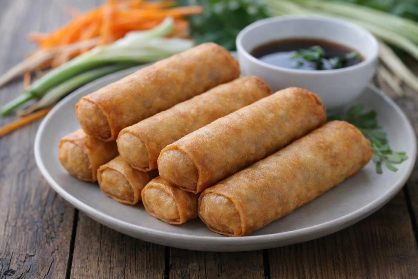 A plate of golden brown bean sprout and vegetable spring rolls with fresh vegetables and a bowl of dipping sauce on a table.