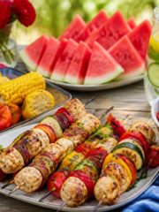 A table outdoors with grilled vegetables, mixed berry salad, chicken and vegetable skewers, watermelon slices, and a pitcher of infused water, set for a healthy Fourth of July meal.